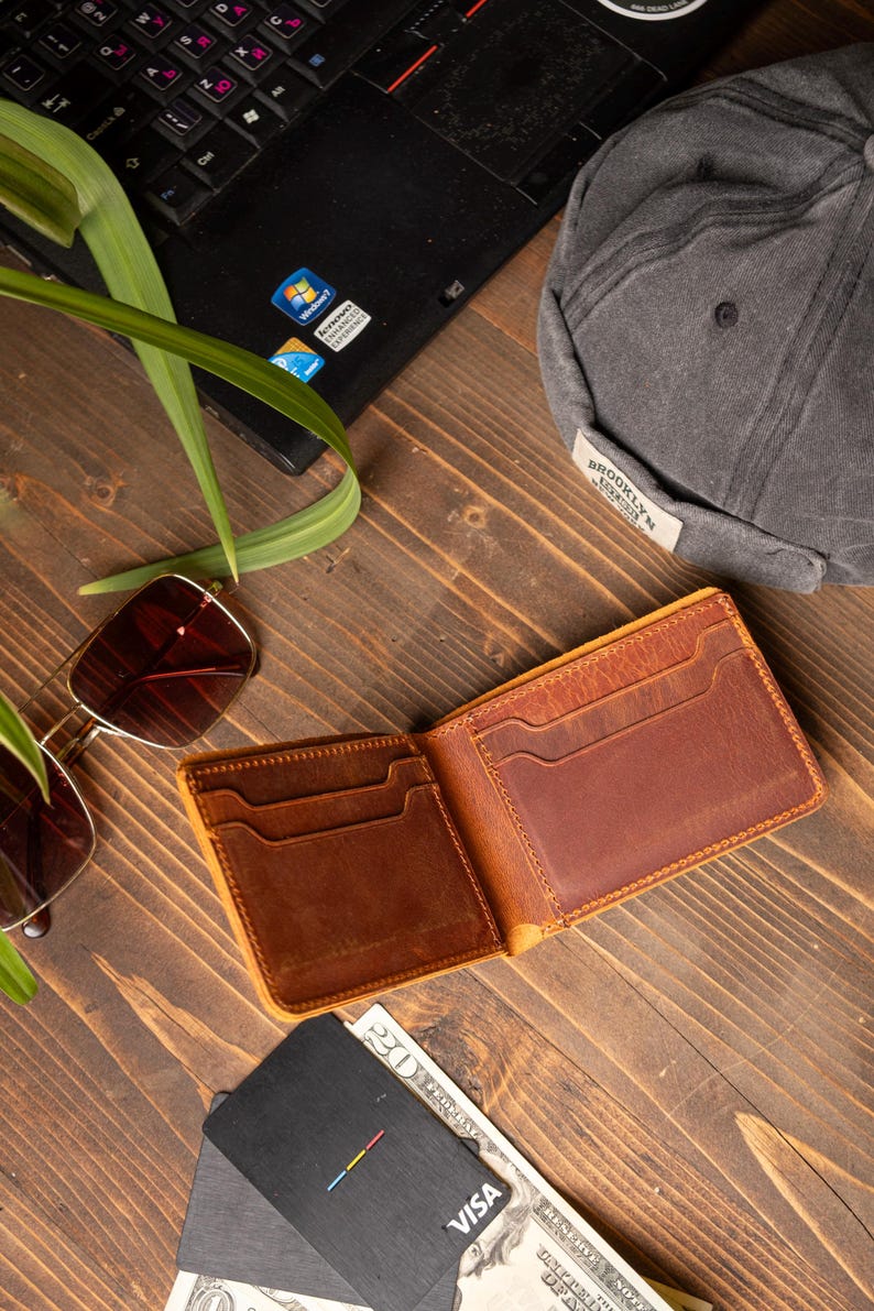 May include: Open, brown leather wallet with multiple card slots, resting on a wooden surface. A pair of sunglasses, a gray cap, a laptop, and a Visa card are also visible. The wallet is empty.