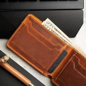May include: Open brown leather wallet with cash and a black card inside. A wooden pen and a black notepad are next to the wallet. A black keyboard is in the background.