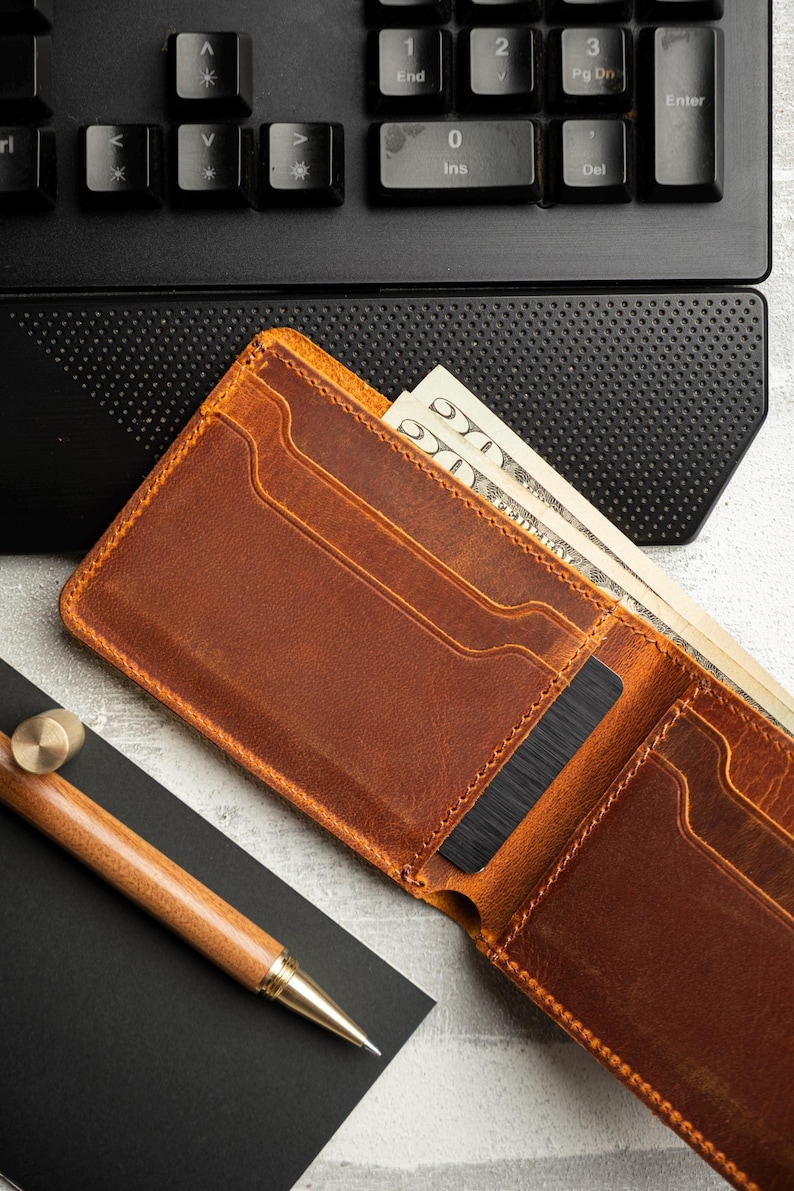 May include: Open brown leather wallet with cash and a black card inside. A wooden pen and a black notepad are next to the wallet. A black keyboard is in the background.