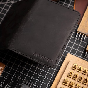 May include: Black leather wallet with the name "ROBERT" embossed on the front. The wallet is open, revealing the interior. Crafting tools, including needles, thread spools, and letter stamps, surround the wallet.