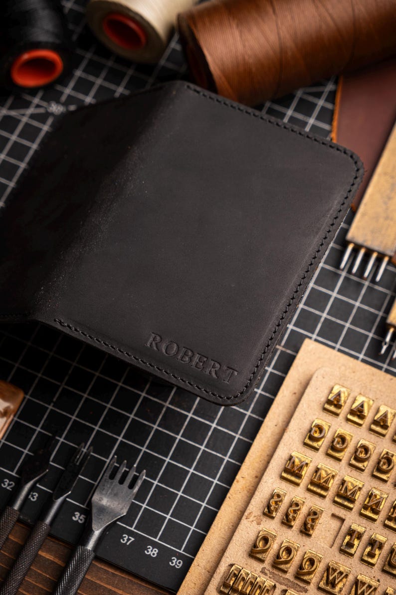 May include: Black leather wallet with the name "ROBERT" embossed on the front. The wallet is open, revealing the interior. Crafting tools, including needles, thread spools, and letter stamps, surround the wallet.