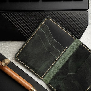 May include: A dark green leather wallet with multiple card slots, cream stitching, and a matching green interior. A wooden pen with gold accents and a black notebook are next to the wallet. A black keyboard is in the background.