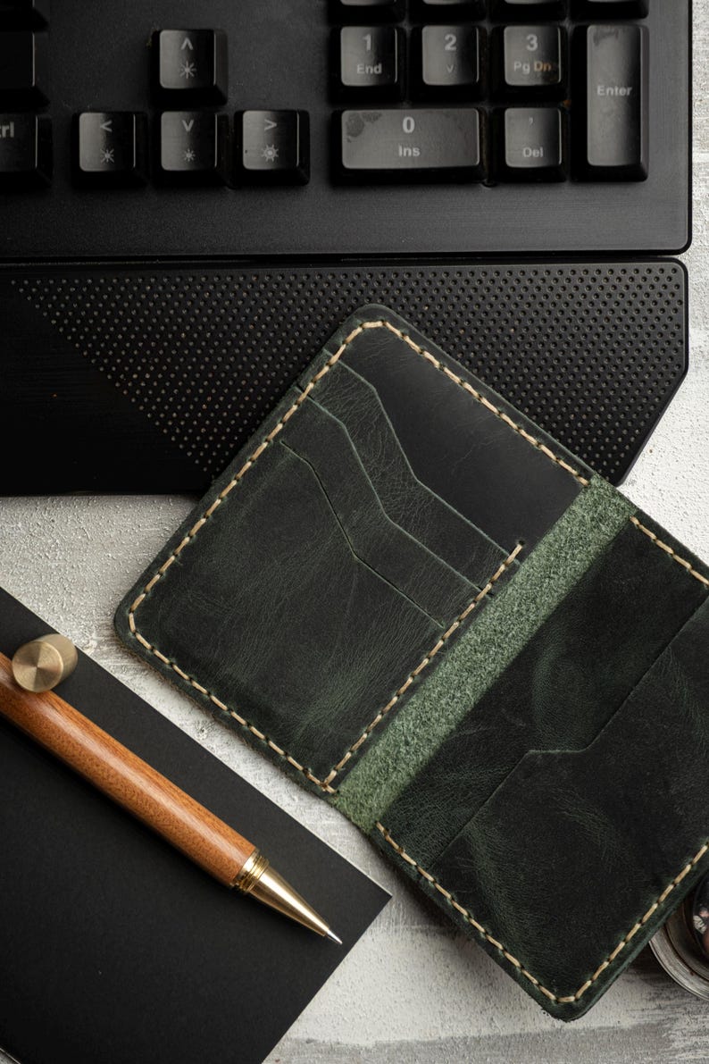 May include: A dark green leather wallet with multiple card slots, cream stitching, and a matching green interior. A wooden pen with gold accents and a black notebook are next to the wallet. A black keyboard is in the background.