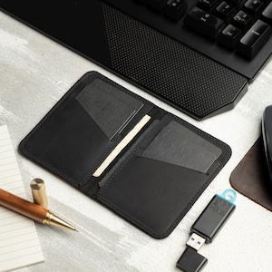 May include: Open black leather wallet with card slots, resting on a light gray surface. A wooden pen, notepad, and a USB drive are also visible. A black keyboard is in the background.