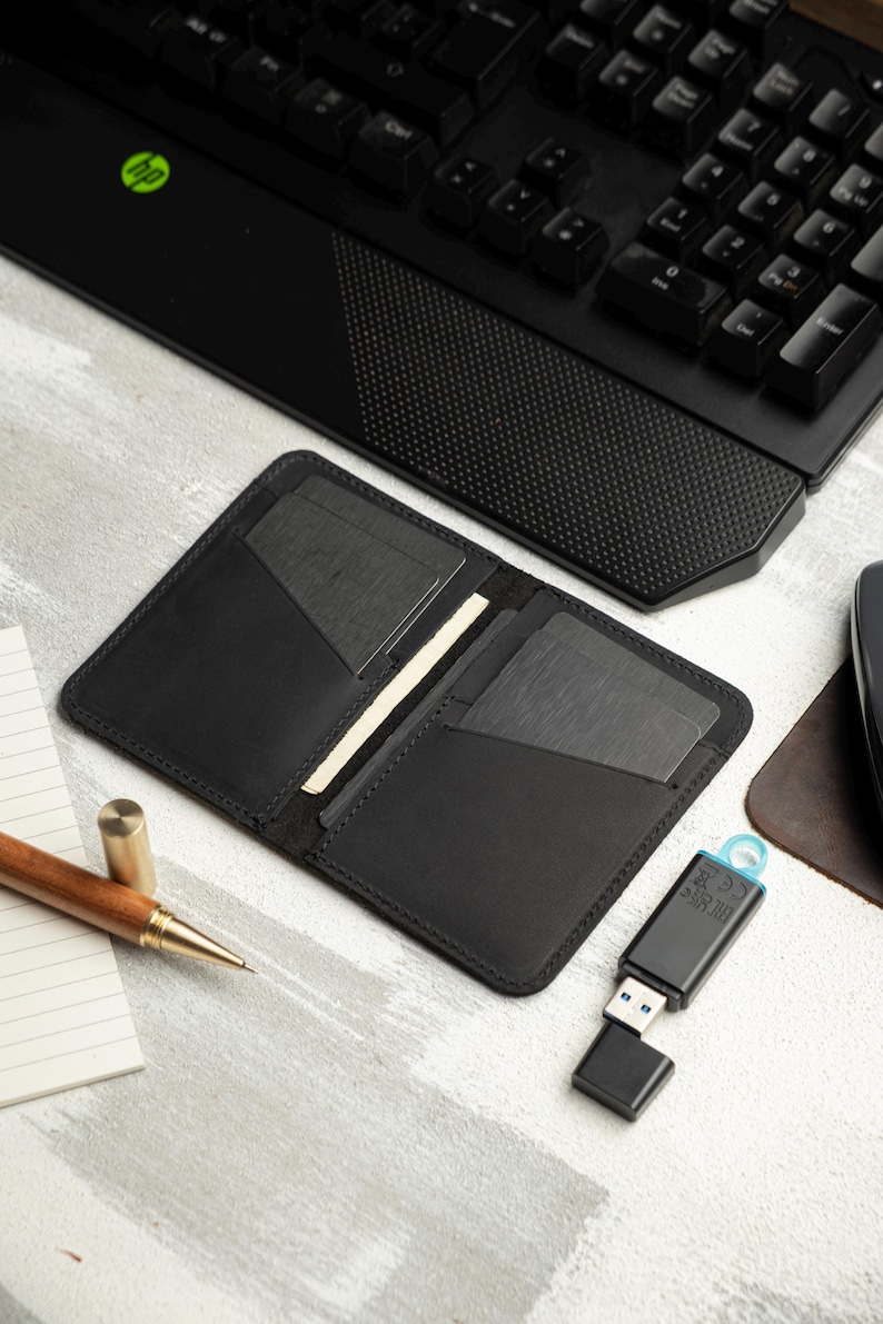 May include: Open black leather wallet with card slots, resting on a light gray surface. A wooden pen, notepad, and a USB drive are also visible. A black keyboard is in the background.