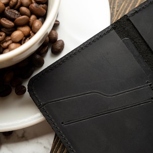 May include: A black leather wallet with card slots is displayed next to a white coffee cup filled with roasted coffee beans. The wallet has a stitched border and a minimalist design, suggesting a focus on functionality and style.