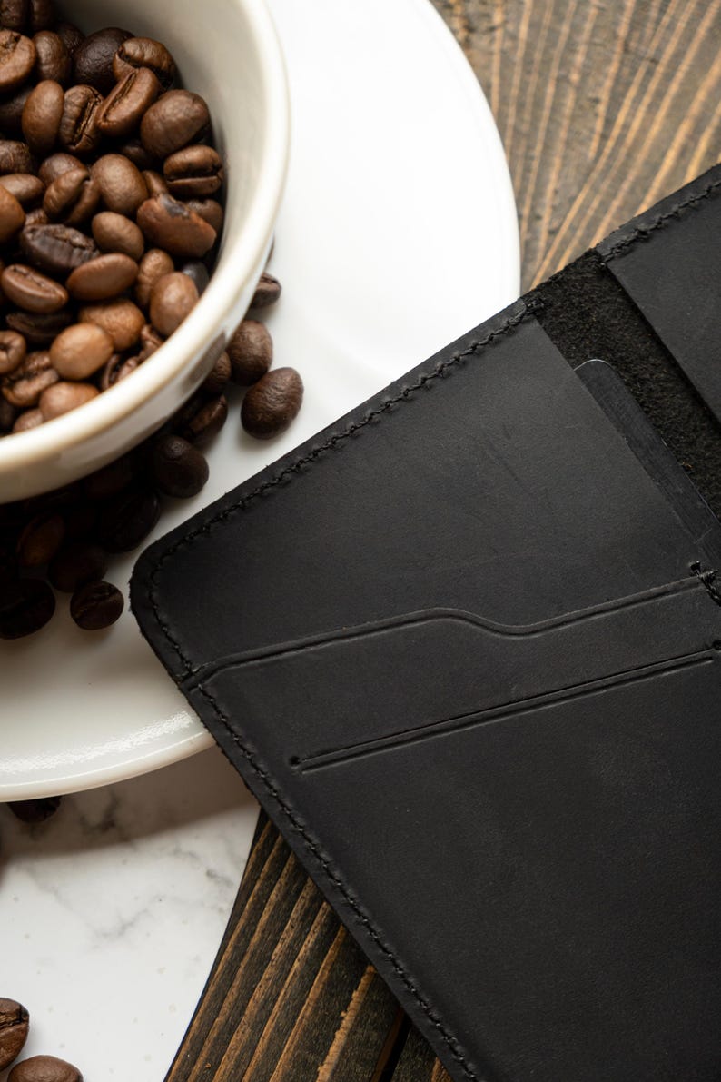 May include: A black leather wallet with card slots is displayed next to a white coffee cup filled with roasted coffee beans. The wallet has a stitched border and a minimalist design, suggesting a focus on functionality and style.