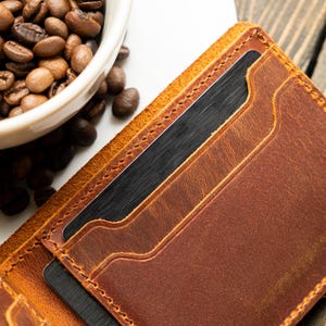 May include: A close-up of a brown leather wallet with multiple card slots. The wallet is open, revealing dark cards inside. A white bowl filled with coffee beans sits in the background, suggesting a coffee-related theme.