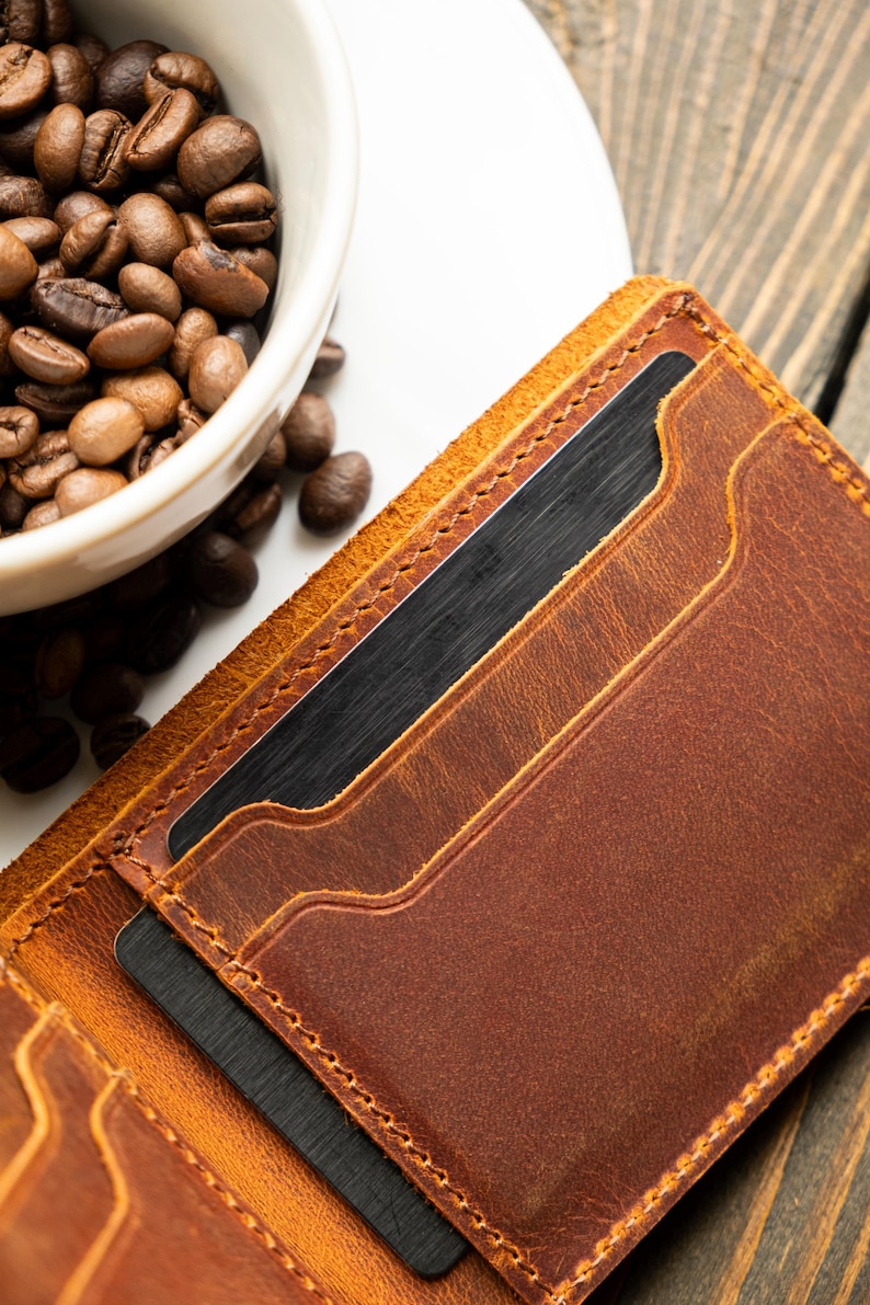 May include: A close-up of a brown leather wallet with multiple card slots. The wallet is open, revealing dark cards inside. A white bowl filled with coffee beans sits in the background, suggesting a coffee-related theme.