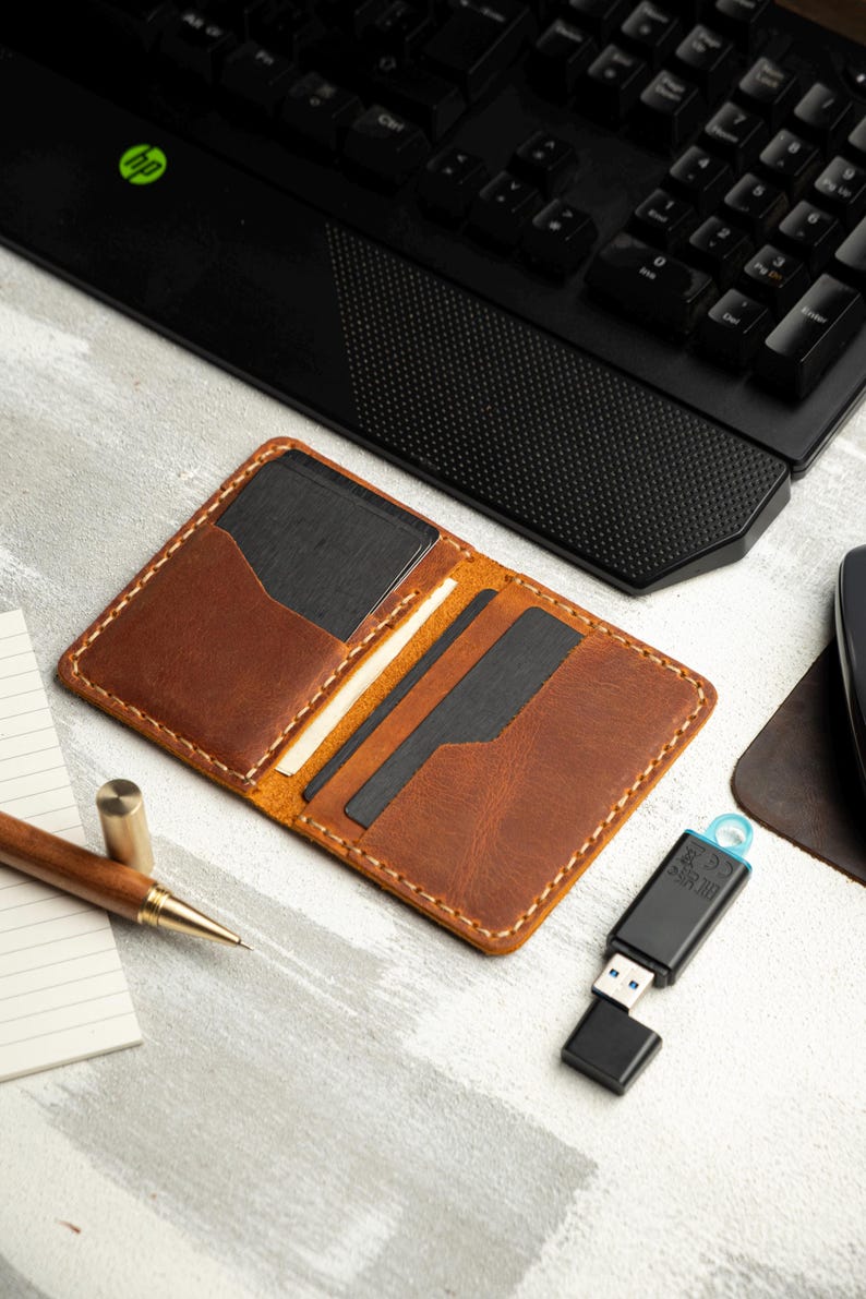May include: Open brown leather wallet with multiple card slots, a black interior, and white stitching. A USB drive and a wooden pen are also visible on a light-colored surface, along with a black keyboard.