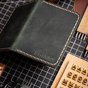 May include: Dark green leather wallet with the name "ROBERT" embossed on the front. The wallet has cream-colored stitching around the edges. Crafting tools and spools of thread are in the background.