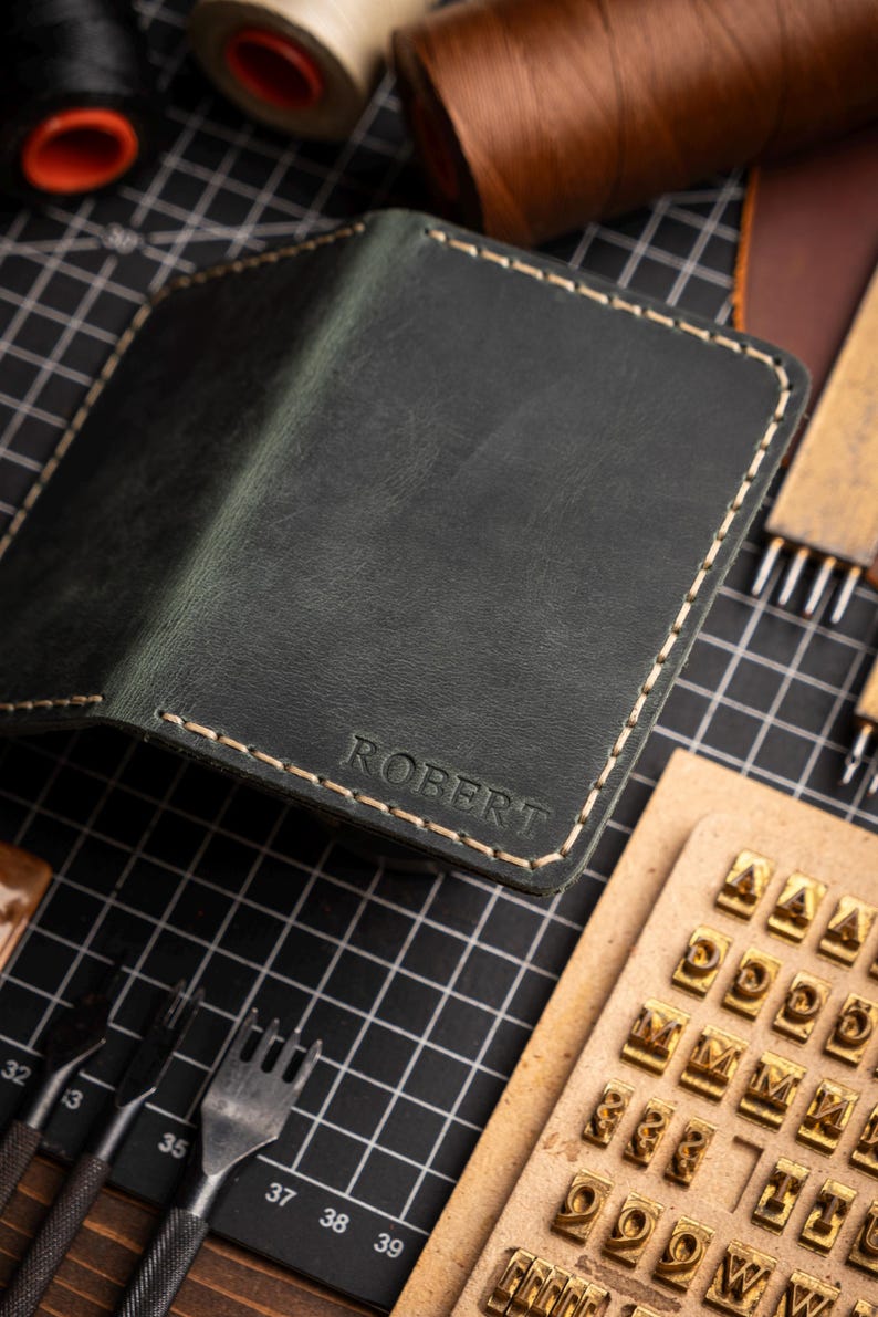 May include: Dark green leather wallet with the name "ROBERT" embossed on the front. The wallet has cream-colored stitching around the edges. Crafting tools and spools of thread are in the background.
