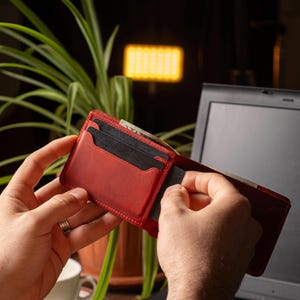 May include: Open red leather wallet with multiple card slots. The wallet is held open, revealing the interior and a few cards. The stitching is visible around the edges. The background includes a laptop, a plant, and a lamp.