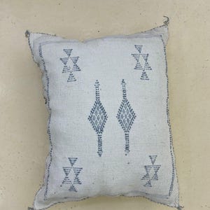 Könnte beinhalten: Ein quadratisches, cremefarbenes Kissen mit einem geometrischen Design in einem gedämpften Blau-Grau. Das Kissen hat Rauten- und Dreiecksmuster mit einem genähten Rand. Das Kissen ist etwa 45 cm groß.