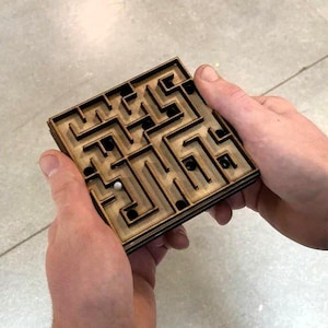 Puede incluir: Un juego de laberinto de madera con una pequeña bola de metal en su interior. El laberinto es de forma cuadrada con caminos intrincados y está sostenido por dos manos. La madera es de color marrón claro.