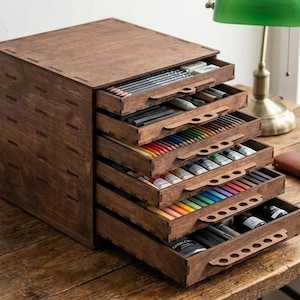 May include: A dark brown wooden storage chest with five pull-out drawers, each featuring a handle with circular cutouts. The drawers are filled with various art supplies, including pencils, colored markers, paint tubes, and small bottles.
