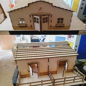 May include: A wooden model of a barn or stable, featuring a pitched roof, small windows, and double doors. The model is constructed from light-colored wood, with intricate details like a horse silhouette and decorative trim. The design includes a fenced area.
