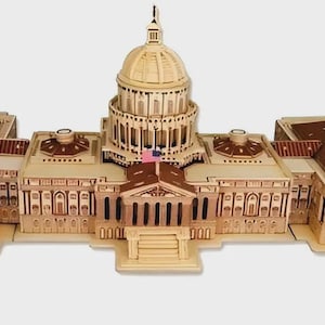 May include: A wooden model of a building with a large central dome and two wings. The model is made of light-colored wood with darker brown accents on the roof. A small American flag is visible.