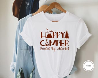 May include: White t-shirt with the words "HAPPY CAMPER Fueled By Alcohol" in brown lettering. The design includes a mountain, teepee, tree, and crescent moon. The shirt is hanging on a wooden hanger.