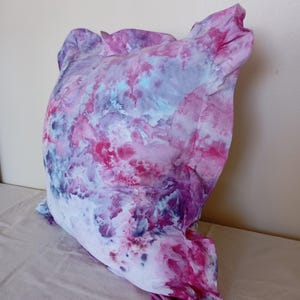 May include: A square throw pillow featuring a tie-dye pattern in shades of magenta, deep purple, and light blue. The fabric has a ruffled edge detail around the perimeter, creating a soft, textured appearance with a marbled, cloud-like color effect.
