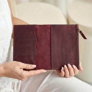 Puede incluir: Una cartera de cuero burdeos con cierre de cremallera y múltiples bolsillos interiores. La cartera está abierta, revelando un interior rojo oscuro contrastante. El diseño es simple y elegante, adecuado para llevar tarjetas y efectivo.