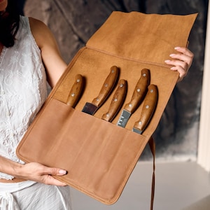 May include: A brown leather knife roll with five knives. The knives have wooden handles and stainless steel blades. The roll is open, displaying the knives in individual slots. The leather is a light brown color.