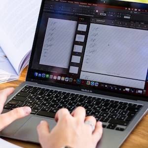 Puede incluir: Un ordenador port&aacute;til MacBook Air que muestra una hoja de pr&aacute;ctica de caligraf&iacute;a en la pantalla. El teclado est&aacute; enfocado, con manos tecleando. Tambi&eacute;n se ve un libro abierto con ilustraciones de margaritas y un tintero de cristal.