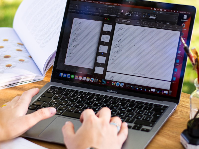 Puede incluir: Un ordenador port&aacute;til MacBook Air que muestra una hoja de pr&aacute;ctica de caligraf&iacute;a en la pantalla. El teclado est&aacute; enfocado, con manos tecleando. Tambi&eacute;n se ve un libro abierto con ilustraciones de margaritas y un tintero de cristal.