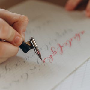 Puede incluir: Primer plano de una mano usando una pluma de caligraf&iacute;a para escribir en papel rayado. La pluma es negra y plateada, y la tinta es de un rosa vibrante. El papel tiene l&iacute;neas tenues y algo de texto preescrito.