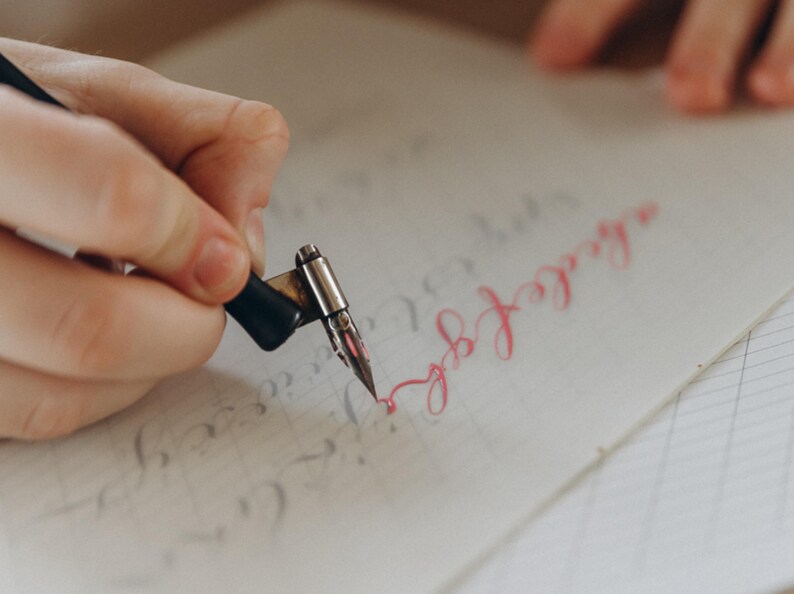 Puede incluir: Primer plano de una mano usando una pluma de caligraf&iacute;a para escribir en papel rayado. La pluma es negra y plateada, y la tinta es de un rosa vibrante. El papel tiene l&iacute;neas tenues y algo de texto preescrito.