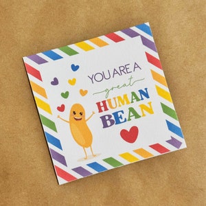 Puede incluir: Una tarjeta blanca con un borde de arcoíris y un frijol de dibujos animados con una cara sonriente. La tarjeta dice "You are a great human bean" con un corazón rojo.