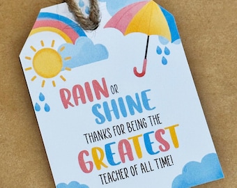 Op de afbeelding: Een blauwe en witte cadeau-tag met een regenboog, zon en paraplu. De tag zegt "Rain or Shine Thanks for being the Greatest Teacher of All Time!"