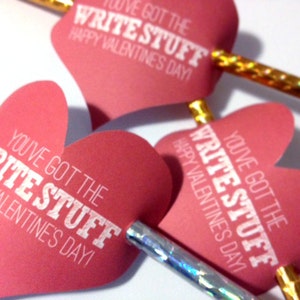 May include: Close-up of Valentine's Day pencils with heart-shaped toppers. The pink hearts have white text that reads "You've Got The Write Stuff Happy Valentine's Day!". The pencils have gold and silver accents.