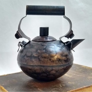 May include: A round, hammered metal teapot with a dark, burnished bronze finish. It features a central lid with a small cylindrical knob, a curved spout, and a high, arched handle attached to the sides with decorative, wavy metal brackets.