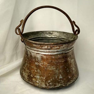 May include: A weathered copper bucket with a wide, open top and a rounded body. The exterior shows a mix of tarnished copper and worn silver-toned finish. It features a curved metal handle attached to two side loops, showing signs of oxidation.