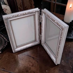 May include: A hinged, silver-colored picture frame with ornate detailing. The frame is open, revealing two clear glass panels. The frame has a decorative border and a hinged design, suitable for displaying two photos or images.