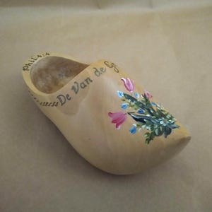 May include: A light-colored wooden clog featuring a hand-painted floral design with pink and blue flowers on the toe. The side of the shoe is inscribed with the text Phil 4:4 and De Van de G in dark lettering, with a hollow interior space.