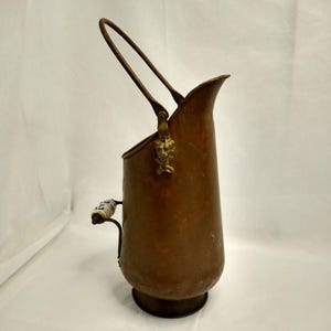 May include: A copper coal scuttle featuring a tall, arched metal handle and a side handle with a white ceramic grip decorated with blue patterns. The vessel has a wide, open top and a brass-colored lion head ornament at the handle attachment point.