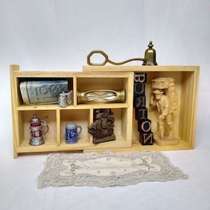 Może przedstawiać: Drewniana półka ekspozycyjna z różnymi przedmiotami w stylu vintage. Zawiera miniaturową klepsydrę, kufle, figurkę statku i rzeźbioną drewnianą figurkę. Słowo "BURTON" jest zapisane czarnymi klockami literowymi. Na dole znajduje się dekoracyjna koronkowa serwetka.