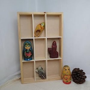 May include: A light-colored wooden display shelf with nine rectangular compartments. Inside are a yellow wooden shoe, a blue nesting doll, a dark red figurine, and a small metal object. A second nesting doll and a pinecone sit in front of the shelf.