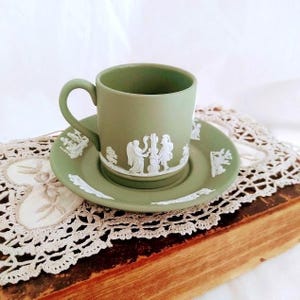 May include: A sage green teacup and saucer set. The cup and saucer feature white bas-relief classical figures and decorative elements. The set is displayed on a vintage book and lace doily.