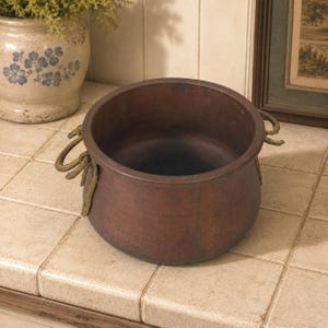May include: A round, dark brown metal pot with a wide opening and a smooth, matte finish. The pot features two textured, brass-colored metal handles attached to opposite sides, each with a curved, organic shape and a slightly weathered appearance.