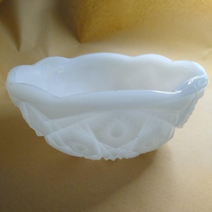 May include: A white milk glass bowl featuring a scalloped rim and a textured, pressed pattern on the exterior. The bowl has a translucent quality and a rounded, shallow shape with a decorative design that includes geometric and floral motifs.