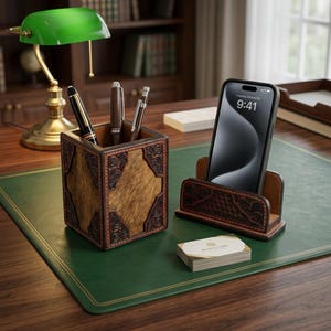 May include: A square brown leather pen holder with embossed floral corners containing three pens, alongside a matching leather smartphone stand holding a black phone displaying 9:41, and a small stack of white business cards with gold accents.