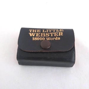 May include: A small, black leather case with a flap and button closure. The gold lettering on the flap reads "THE LITTLE WEBSTER 18000 Words". The case is rectangular and appears to be a miniature dictionary.