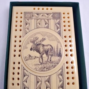 Può includere: Tavola da cribbage color crema con un'illustrazione dettagliata di un alce in una cornice circolare. La tavola presenta motivi geometrici decorativi e numerosi fori per pioli. La tavola è in una scatola verde scuro.