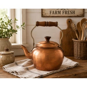 May include: A polished copper tea kettle with a rounded body, a curved spout, and a hinged handle featuring a dark brown wooden grip. The lid is topped with a matching dark brown wooden knob, and the metal frame connecting the handle is brass.