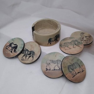 May include: A set of ceramic coasters with a matching holder. Each coaster features a different African animal illustration in black and white, including a zebra, leopard, and rhino. The holder and coasters have a neutral color palette.