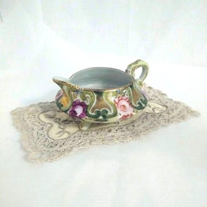 May include: A porcelain creamer pitcher featuring a colorful floral pattern with pink and purple roses, gold-toned scrollwork, and green accents. The vessel has a curved handle, a small spout, and three short feet, resting on a rectangular lace doily.