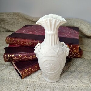 May include: A white, ornate vase with a ruffled top and two handles. The vase features a detailed floral and geometric design. The vase is placed in front of a stack of books with burgundy covers and gold accents.